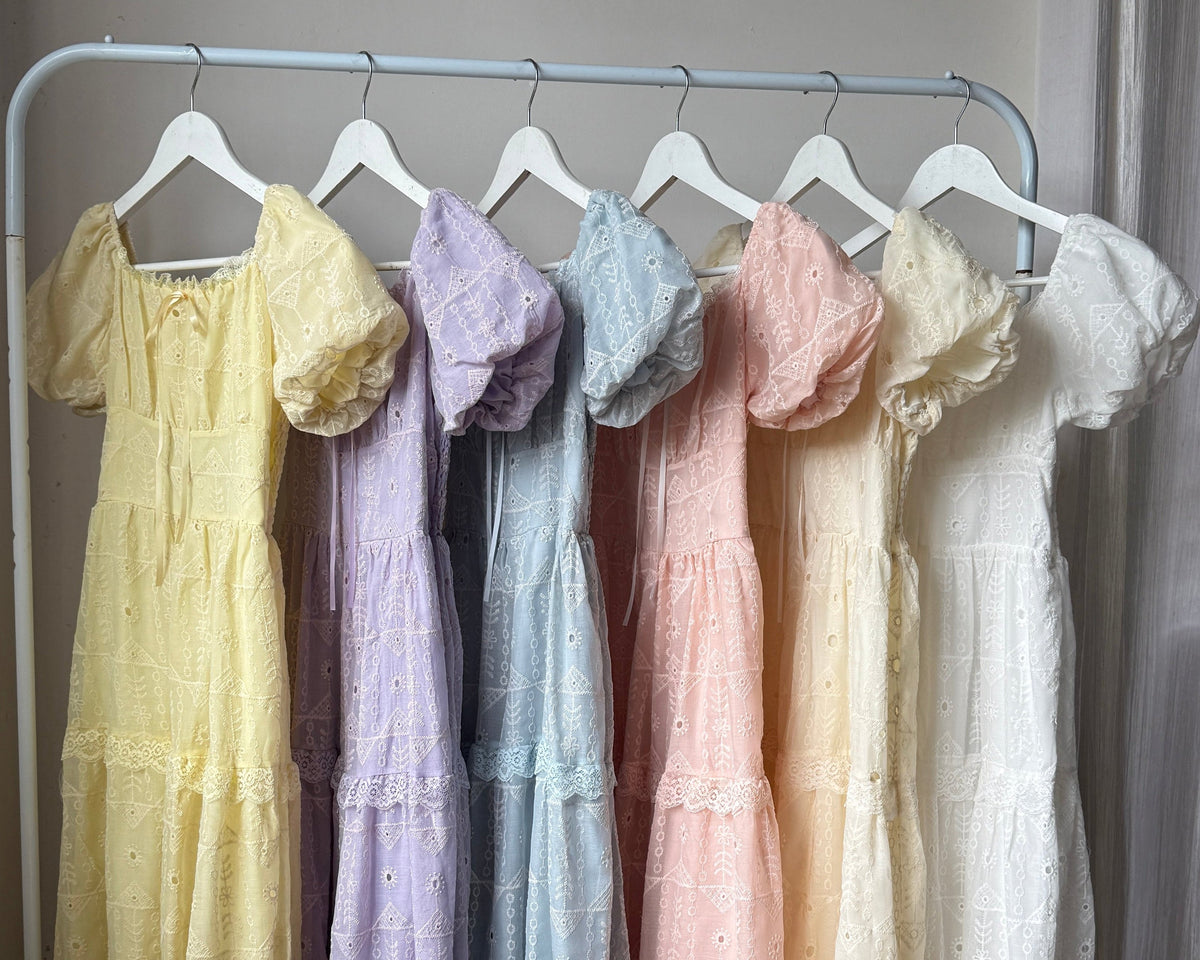 Six tiered cotton dresses in pastel shades of yellow, lavender, light blue, pink, cream, and white, hanging on a white clothing rack in a room with wooden floors and a curtain.