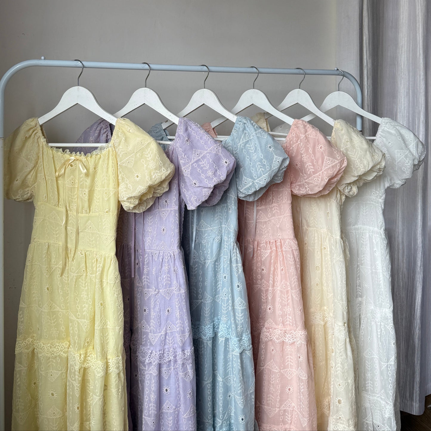 Six pastel-colored eyelet lace dresses in yellow, purple, blue, pink, cream, and white are displayed on a white clothing rack against an indoor wall.