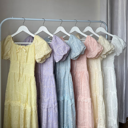 Six pastel-colored eyelet lace dresses in yellow, purple, blue, pink, cream, and white are displayed on a white clothing rack against an indoor wall.