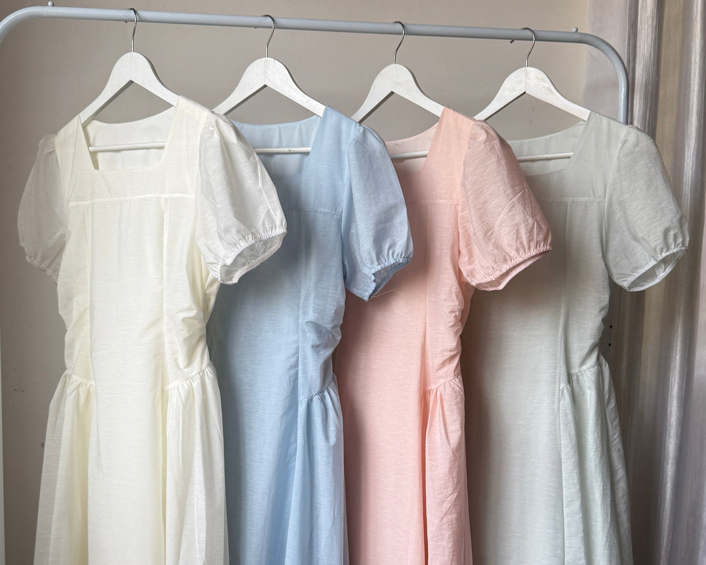Four short puff-sleeve midi dresses in cream, light blue, blush pink, and pale sage green hanging on a white clothing rack in an indoor room with wood flooring and curtains.